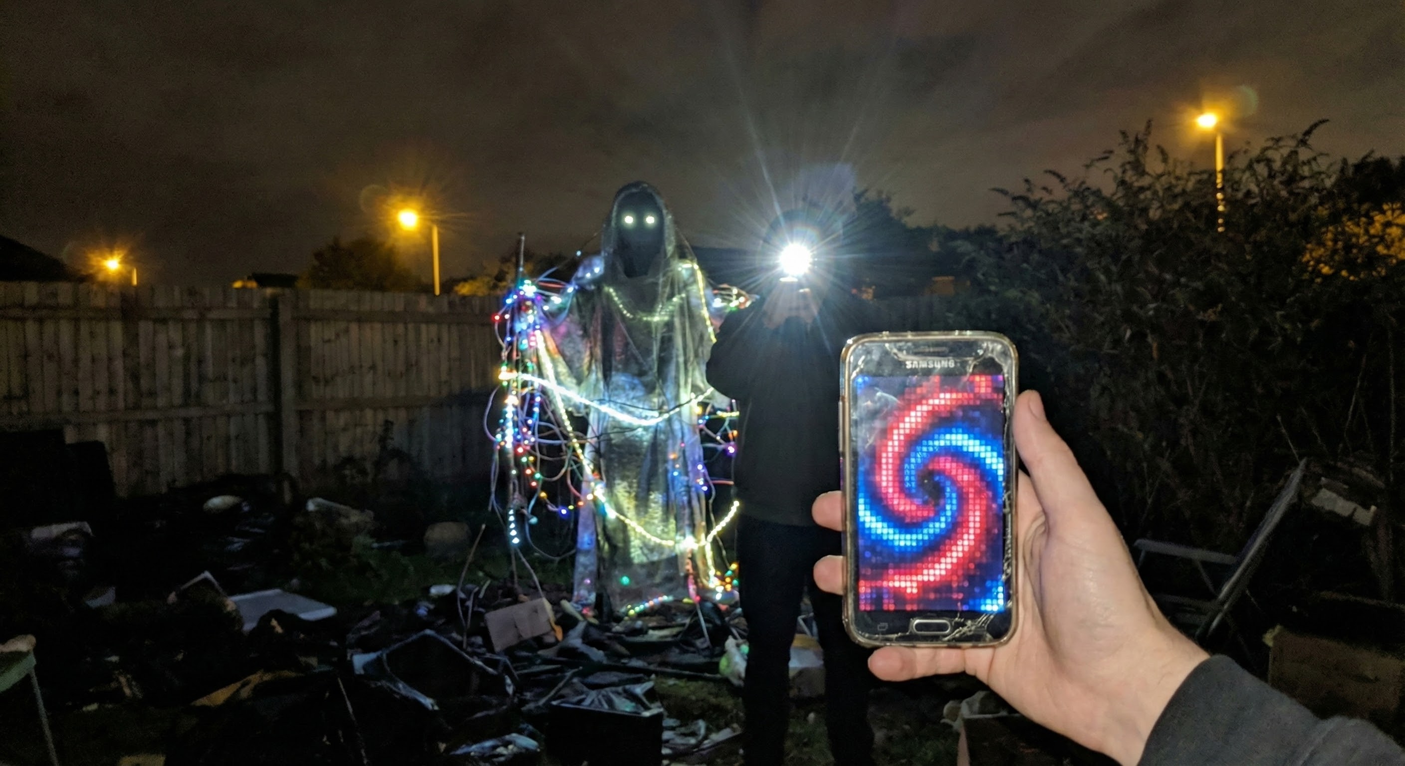 A ghostly figure with glowing eyes stands in a cluttered backyard at night, draped in translucent fabric and tangled Christmas lights, while a hand holds up a phone displaying a hypnotic red and blue spiral