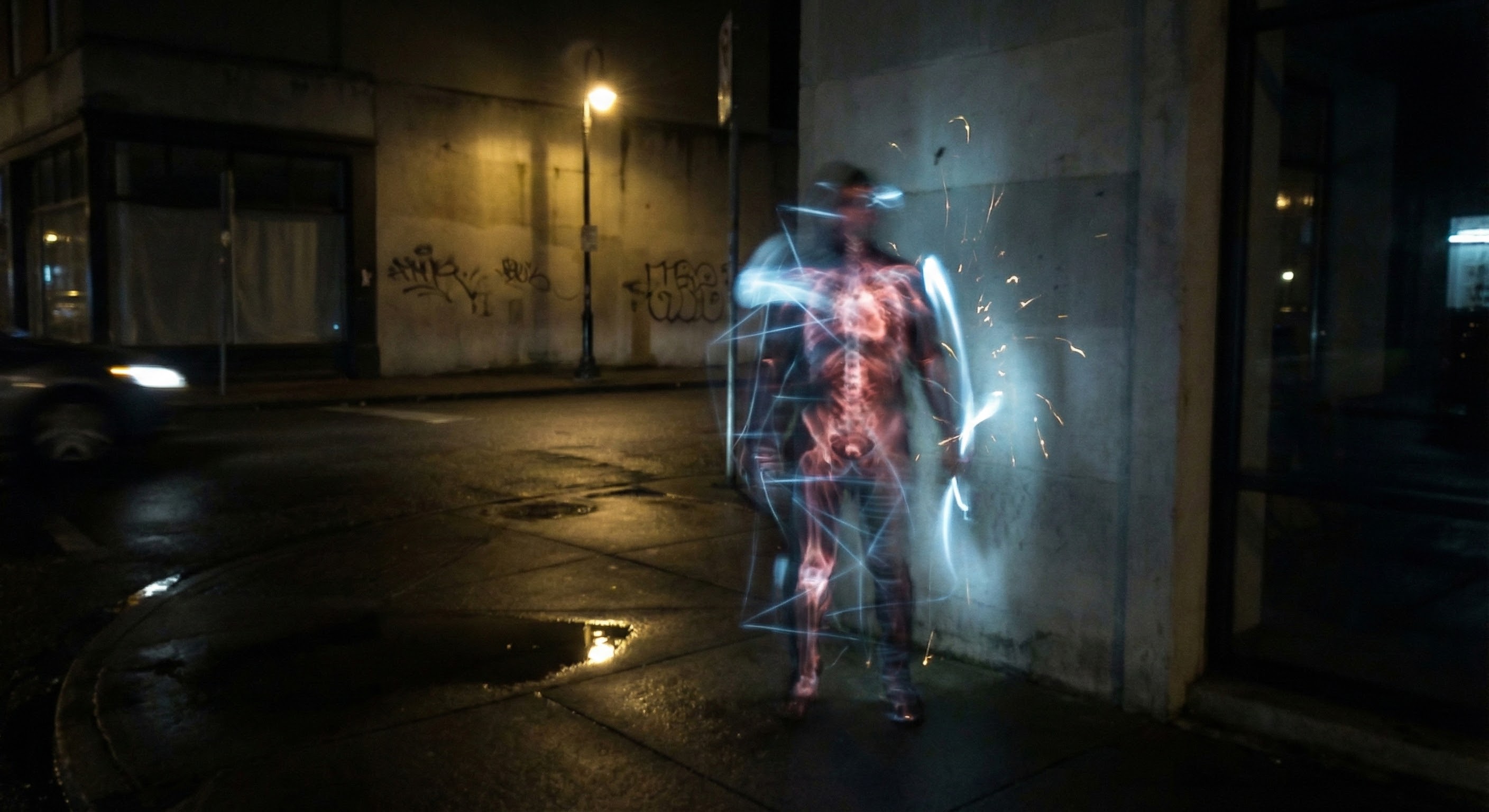 Long exposure of a figure on a wet urban street corner at night, their body blurred and ghostly with a glowing anatomical skeleton visible through them, surrounded by light trails and sparks