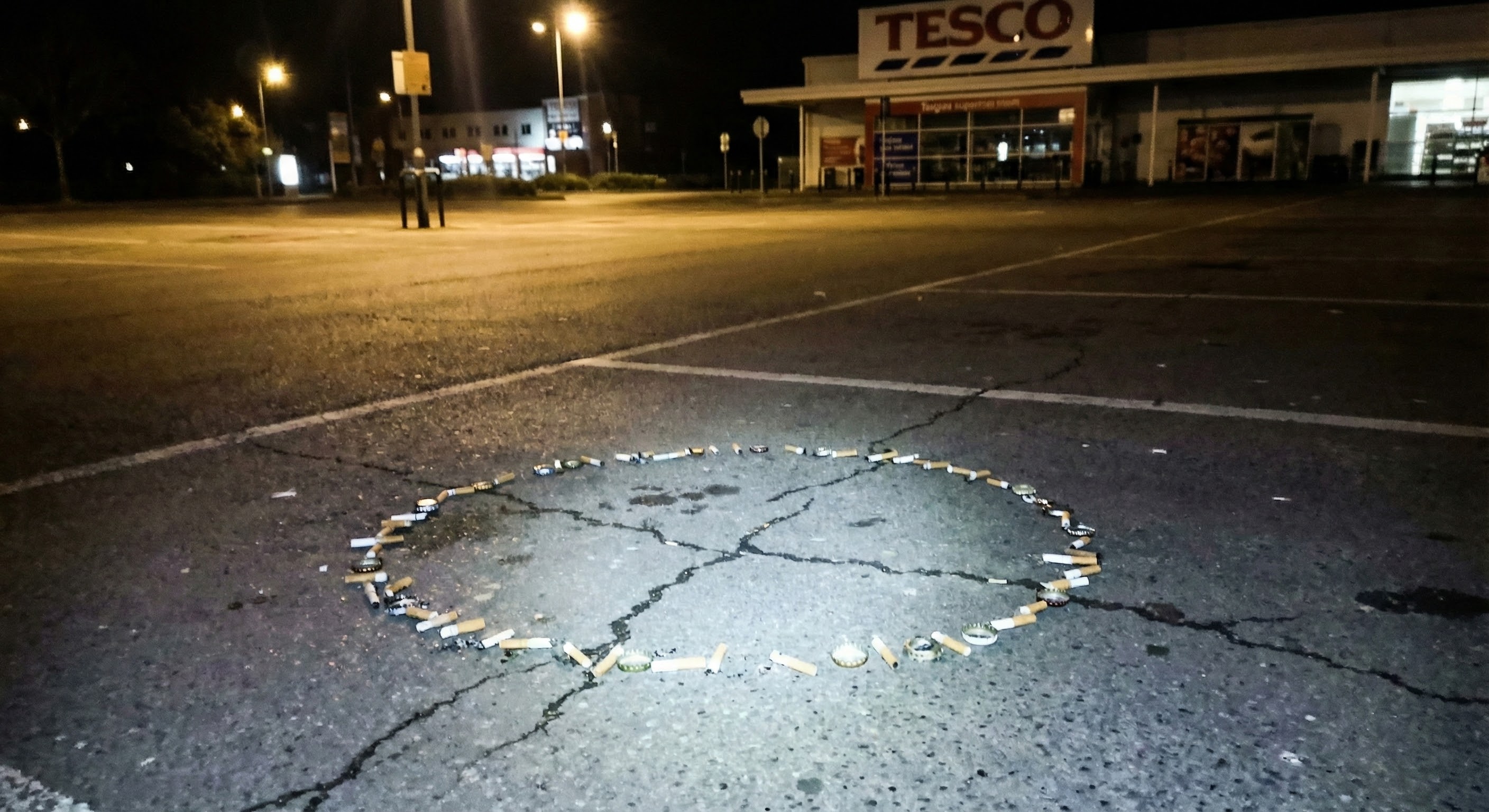 Cigarette butts and bottle caps arranged in a rough circle on cracked tarmac in an empty Tesco car park at night