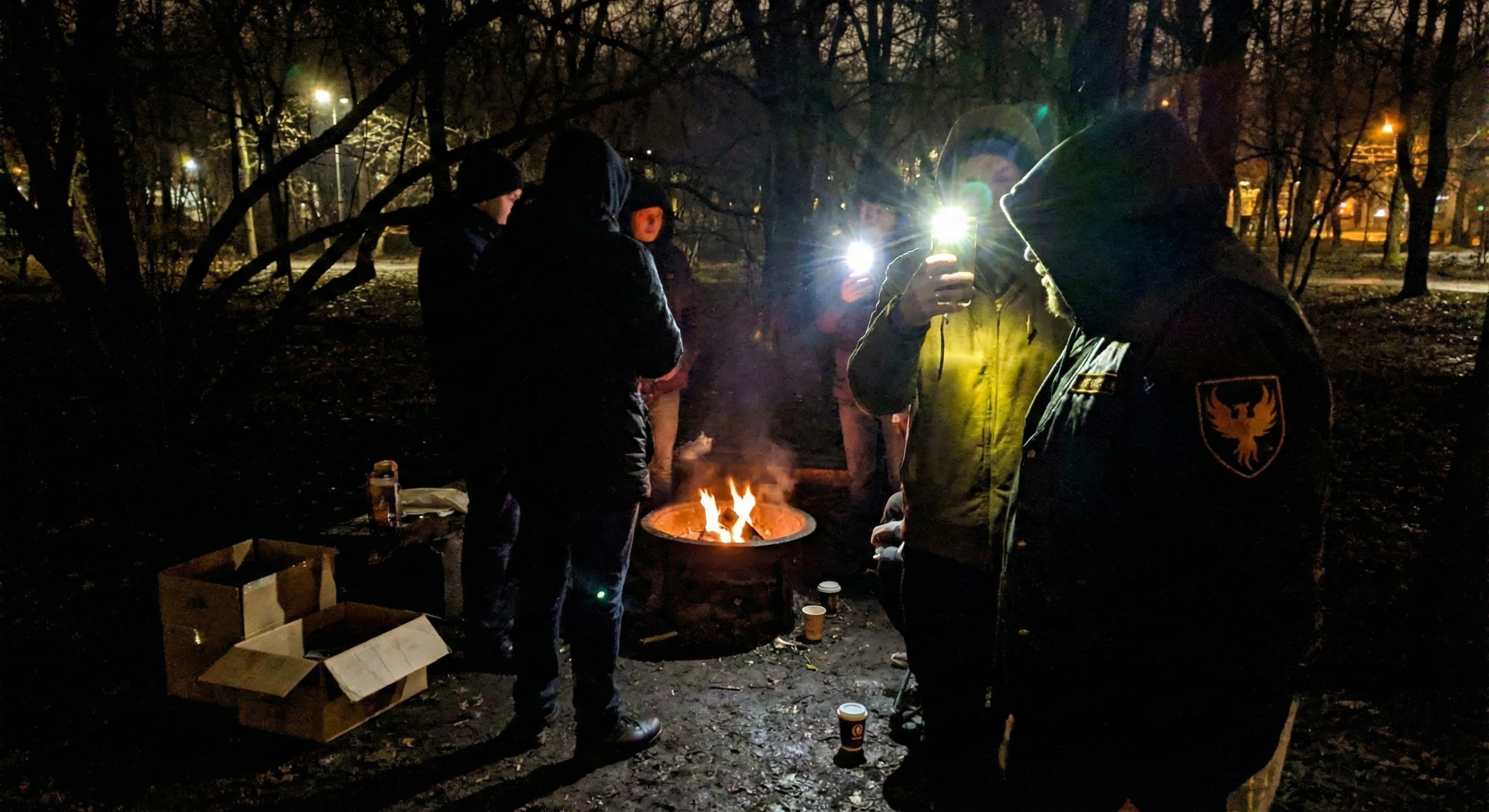 A group of people in winter coats huddle around a fire burning in a metal drum in a dark park, one checking their phone, bare trees and cardboard boxes visible nearby