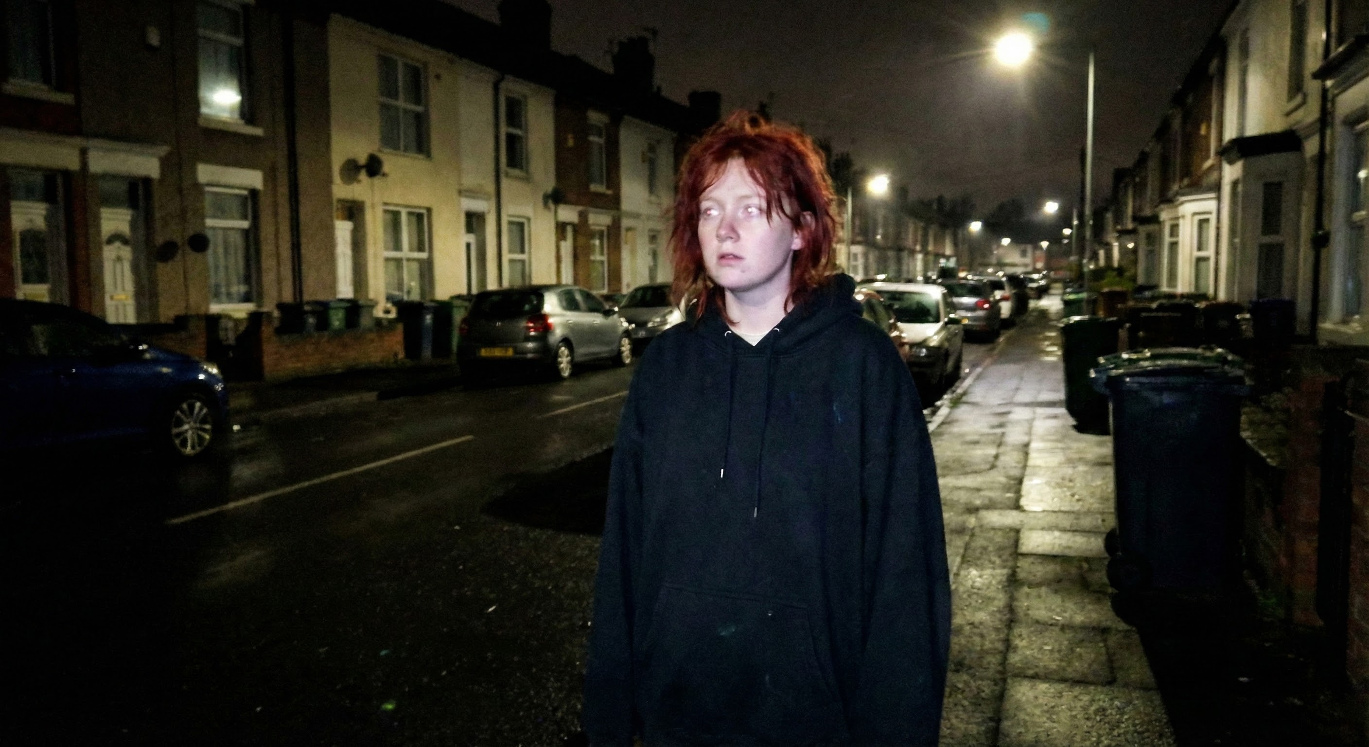 A young person with tousled red hair stands alone on a wet terraced street at night, wearing an oversized black hoodie, gazing upward with an unsettling stillness